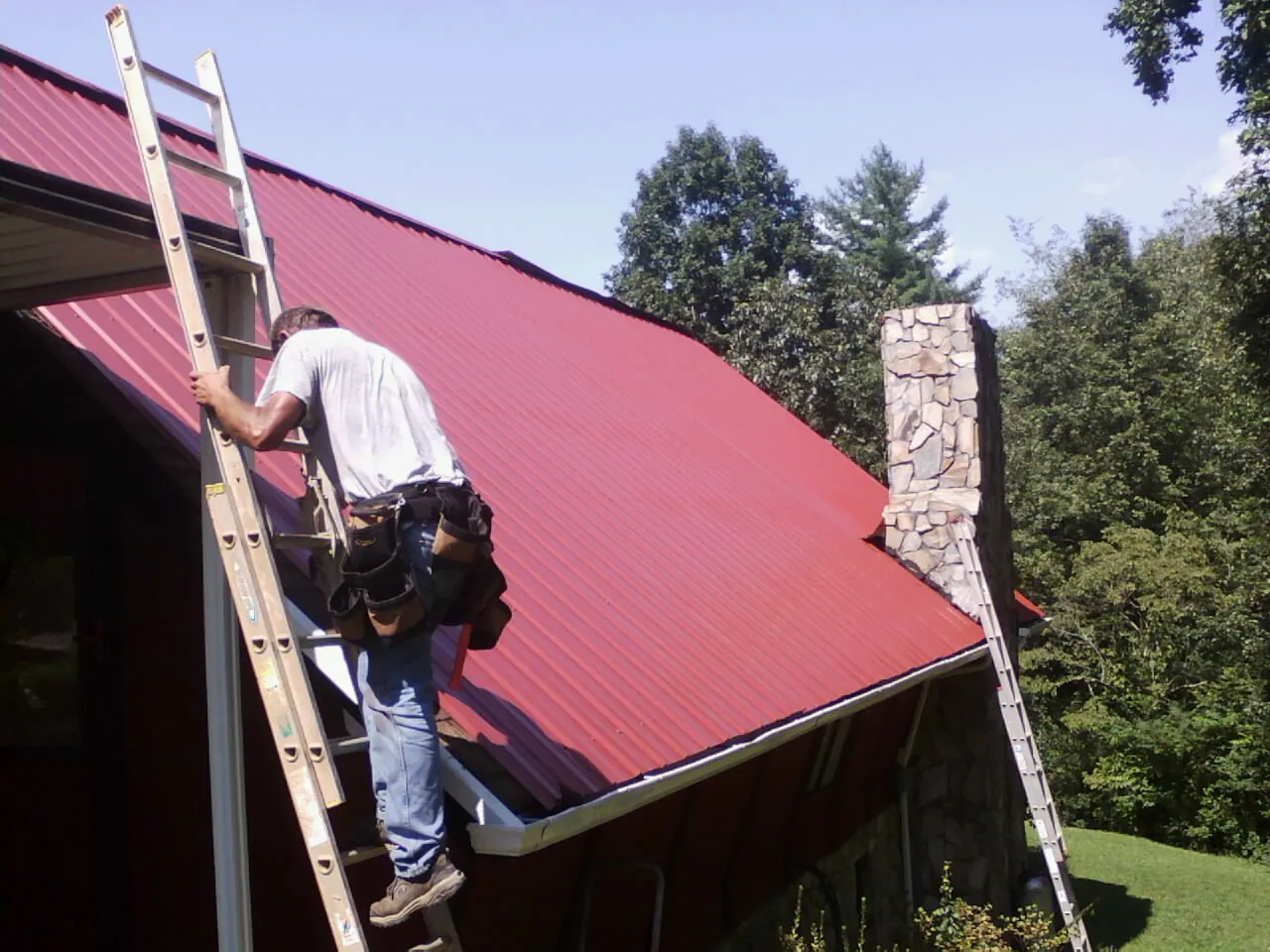 Roofer on a ladder beside a red metal roof and stone chimney — Soffit & Fascia Repair by Roofing Brothers in Jamison